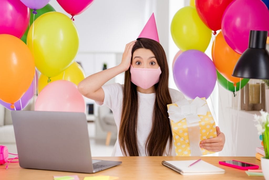 Photo of pupil small school lady notebook online lesson sit table, safety face mask hold big giftbox birthday girl unexpected classmates surprise home party greeting quarantine indoors