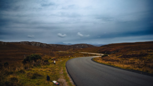 A winding asphalt road curves through a vast, rugged, brown moorland under a dark, dramatic, stormy sky.